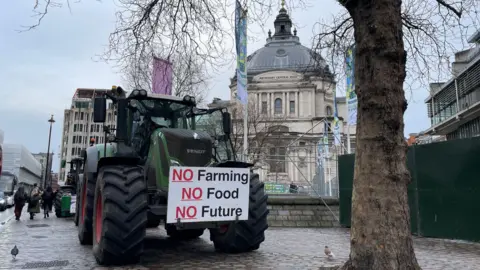 Traktor Malcolm Prior/BBC diparkir di luar Pusat Konferensi di jalan London yang basah. Traktor timah memiliki plakat yang mengatakan "Tidak ada pertanian, tidak ada makanan, tidak ada masa depan"