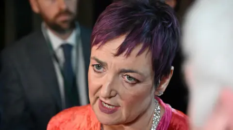 Getty Images Head shot of Angela Constance speaking to journalists in the lobby of the Scottish Parliament. She is wearing a diamond necklace and a bright pink/orange dress.