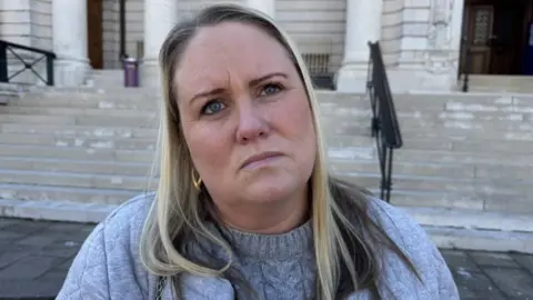 A woman with blonde hair, wearing a grey jumper standing in from of the court steps