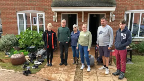 Six residents standing outside their newbuild homes. They are looking unhappily at the camera. A number of random possessions like trainers and plant pots are scattered around their feet. 