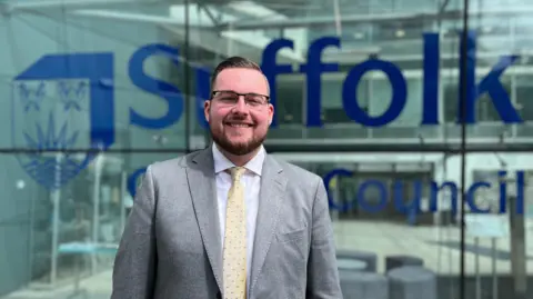 Michael Hadwen is standing in front of a glass building at Suffolk county council. He is wearing a grey suit, white shirt and yellow tie.