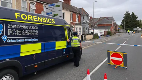 BBC A navy blue police forensics van waits at a police tape cordon on a residential street. The van has forensics written in yellow reflective writing and South West Police Forensics written in blue writing on a black tinted window. An officer in a hi-vis vest stands at the window of the van