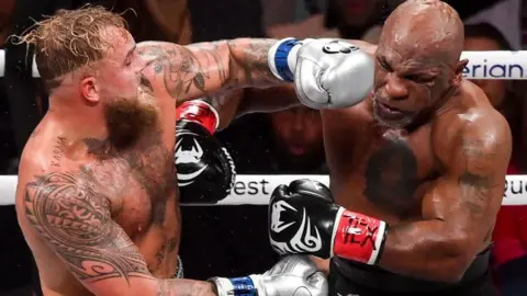 Getty Images Jake Paul, to the left of the frame, lands a punch on Mike Tyson during their so-called boxing match. The event was Netflix's first foray into live sport. Paul is wearing sliver gloves and Tyson's gloves are black with white markings on them, similar to his infamous 'tribal' facial tattoo.