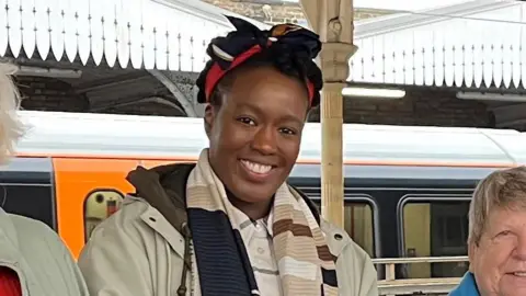 Dalia James is seen from the chest up in smiling in a group picture with others just out of frame to her side on a train platform.