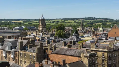 n image of Oxford's skyline with the city in the foreground and rolling hills in the background. The sky is blue.