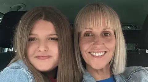 Grace Keeling and Sarah Keeling looking at the camera while sat in a car. Grace has long brown hair and Sarah has long blond hair, both are wearing blue denim jackets.