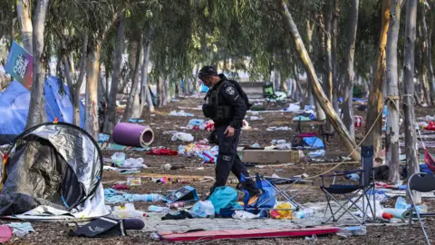 EPA An Israeli police officer walks through the Nova festival site after the attack, where, among the slender trees providing shade, tents are scattered around and personal possessions litter the floor after people fled. On one tree, a sign says, "Chill Out Zone".