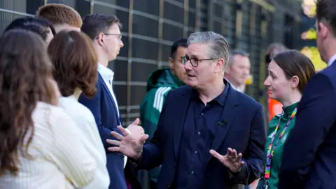 PA Keir Starmer is in a navy suit talking to a small group of people next to a fence. He is at a football pitch and the man standing behind him and woman to his right are wearing green kits. 