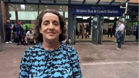 A woman with shoulder-length, curly brown hair poses for a head and shoulders photograph. She is smiling and wearing a black and blue patterned top. She is in front of Bristol Bus & Coach Station, with people seen milling about in the background.