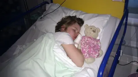 PA Images Jamilla Blake as a seven-year-old asleep on her side in a hospital bed with a teddy bear on the pillow beside her