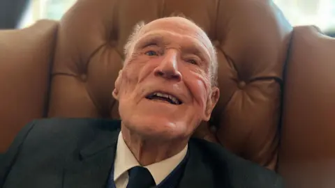 A close up photograph of Len Chivers, wearing a suit and tie while sat on a brown leather chair.