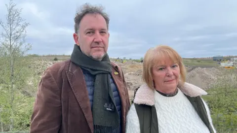 Chris Green and Liz Reid standing near piles of rubble at the quarry site. Chris has short grey hair, a brown tweed jacket and scarf and a blue jumper. Liz has short blonde hair with fringe and wears a white jumper and a black body warmer.