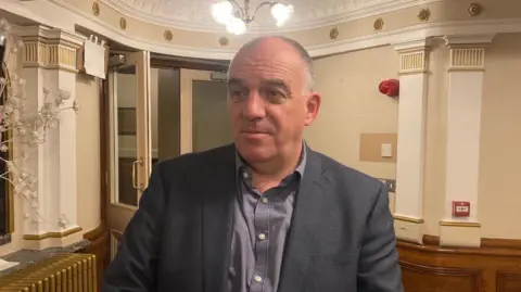 A short crop-haired man in a grey suit and checked shirt