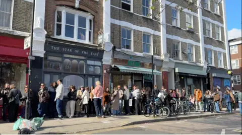 BBC/Rosa Rahimi A large queue outside Logma in Hackney, it is a sunny day and a lot of people are standing and waiting outside a row of shops.