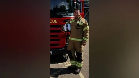 Essex County Fire and Rescue Service A man standing next to a fire engine with a suit on and a helmet under his arm