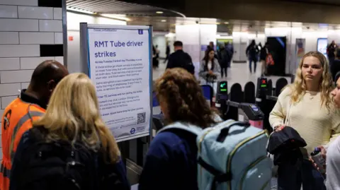 EPA A group of people with bags speak to a TfL worker in an orange high-visibility jacket. They stand in front of a sign that states: RMT Tube driver strikes.