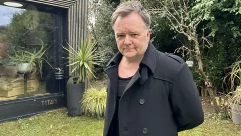 BBC Jason Kerrigan in his garden. There is a lawn and trees behind him. He is wearing a black, low-cut top and a black jacket.  