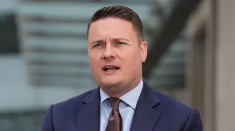Wes Streeting is dressed in a blue suit and brown tie. He has combed, brown hair which is shaved at the sides.