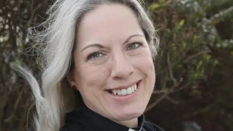Diocese of Portsmouth A woman with long, blonde hair smiles at the camera. She is wearing a clerical collar and is in front of a tree. 