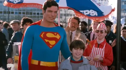 The Cannon Group, Inc/Golan-Globus Productions/Warner Bros A still from Superman IV shows the location filming in Milton Keynes. Superman is smiling with his arm around a boy while a hot dog salesman is standing behind, wearing sunglasses.