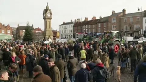 Crowds gathered in the market square. A green tractor is parked on the right.