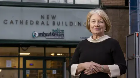 PA Media Marie Anderson. She has a blonde bob, wearing pearl earrings, a black dress with cream cuffs and neckline. She is standing in front of a with a sing on it saying 'New Cathedral Building Police Ombudsman' 