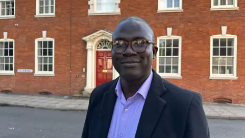 Dr Abiodun Egbetokun wearing a blue suit standing in front of a red brick building