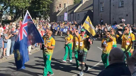 The St Helena team waving their island flag, walking down the streets of Kirkwall with spectators watching and cheering from the side