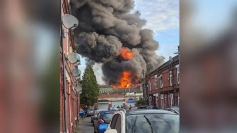 A fiery blaze with orange flames billowing black smoke into the sky over a set of garages at the end of a terraced street.