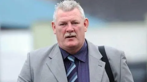 Pacemaker David Tweed goes into the camera. He has short white hair and mustache. He wears a gray blazer, a purple pin with a striped shirt and a tie parked. He carries a bag over his shoulder.