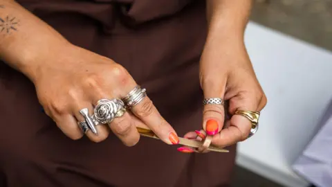 James Mulkeen Image of a woman making a copper ring. Only her hands can be seen, with orange and pink varnished nails. There are elaborate metal rings on most fingers, including her thumb. A copper ring is held between the thumb and index finger of her right hand, and in her left she is holding a file which goes through the ring.