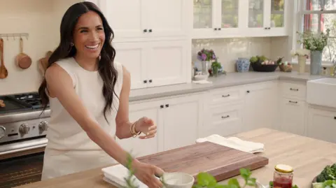 Netflix The Duchess of Sussex pictured wearing a white sleeveless jumper smiling a she places a cup on the kitchen counter