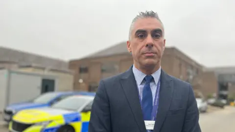 Emma Baugh/BBC Plain clothed detective with grey hair wearing a striped shirt and dark blue suit in front of a police car.