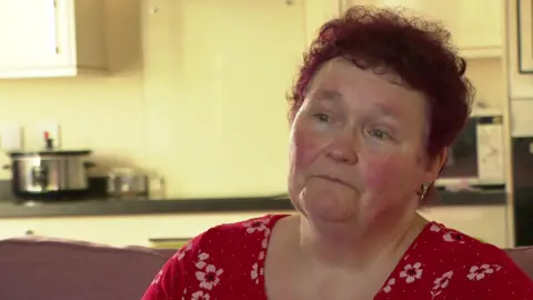 Claire Throssell sits in her flat. She has short, red, curly hair and wears a flowery red top. A kitchen can be seen in the background with a metallic pan and a mircrowave oven.