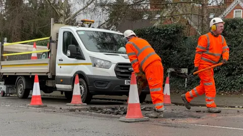 Two men wearing high-vis clothing digging up a road service to repair a pothole. Behind them is a flat-bed van with traffic management equipment on the back. The van and workers are encircled by traffic cones.