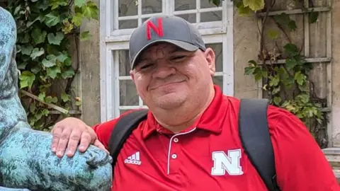 Contributed A man wearing a red polo top with a large white N on it smiles at the camera. He wears a grey cap with a red N on it as well as a backpack. He is resting one hand on a statue next to him.