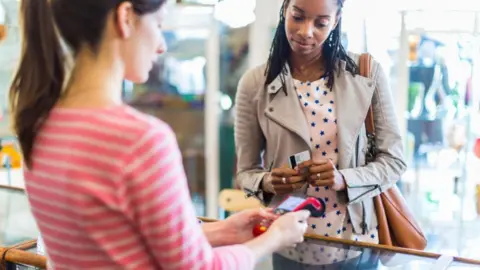 Getty images A woman in a shop holds her bank card ready to pay for an item while a shop worker holds the card machine