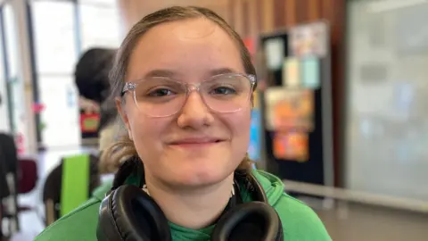 Monica is 17. She has brown hair tied back and transparent plastic spectacles. She wears a green sweatshirt and has headphones around her neck. She is using a wheelchair . 