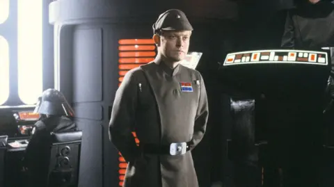 Lucasfilm / Disney Michael Pennington dressed in character as Moff Jerjerrod in Star Wars Return of the Jedi, he wears an imperial robe with a military cap while standing to attention in front of spaceship controls and an unidentifiable pilot.