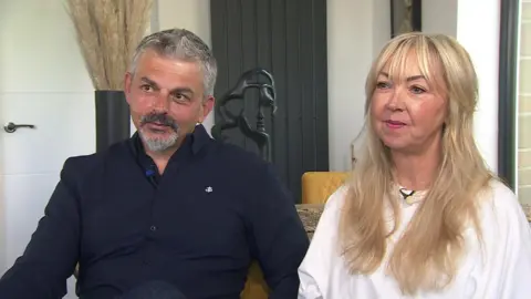 Andrew and Lisa Roussos - he is wearing a blue shirt and has short grey hair, she is wearing a white top and has long blonde hair.