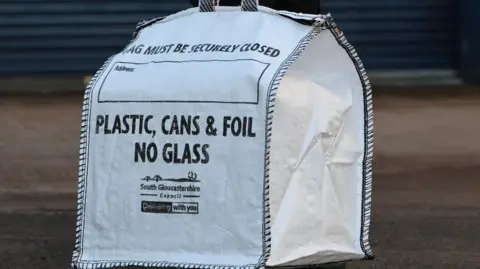 A white heavy-duty bag with "Plastic, Cans and Foil. No Glass" written on the side. In smaller words, South Gloucestershire Council is written.