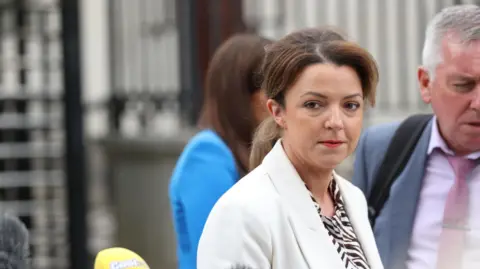PA Media A woman wears a white jacket with a white and black striped top underneath. Beside her, to her left is a man in a grey suit, pink shirt and pink tie. A yellow mic can be seen to her right.