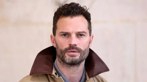 Jamie Dornan is wearing a beige jacket with a brown collar, and looks directly at the camera.
