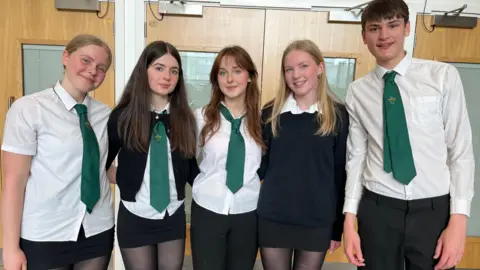Five students in school uniforms standing side by side indoors, wearing white shirts, green ties, and dark skirts or trousers, with wooden doors and frosted glass panels in the background.