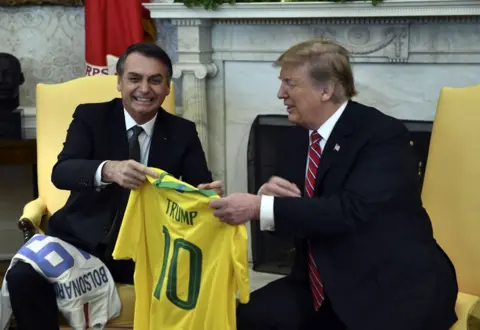 Getty Images Bolsonaro gives Trump a yellow Brazil soccer jersey with the name TRUMP on the back. Trump is smiling as he receives it, seated in the Oval Office.