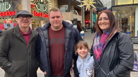 Emma Coombs with her family at Bristol's Cabot Circus. Ms Coombs is wearing a leather look black coat and a pink checked scarf. Her family are all in coats and smiling at the camera. Foot locker is visible in the background.