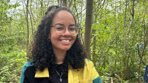 Dr Georgia Kirby smiles at the camera from a forest where she has been monitoring traps. She has long curly dark brown hair and is wearing a cord shirt in shades of green, yellow and black.