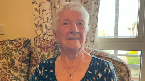 Darren Rozier/BBC An elderly woman with short grey hair sits on a sofa. She is slightly looking up and smiling. She wears a blue blouse with some white spots on it and she wears a gold chain with a cross attached to it around her neck.