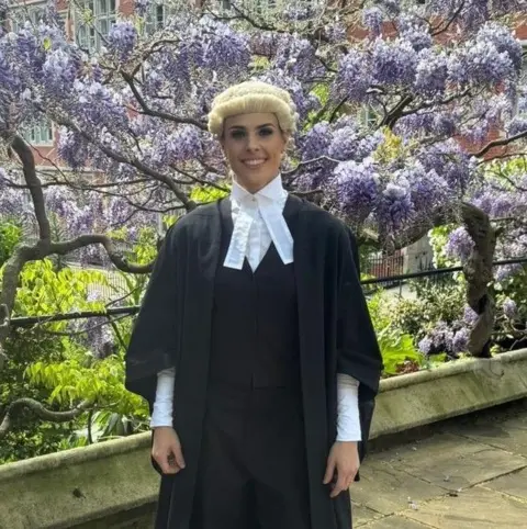 Leonie Hughes A smiling young woman in barrister's clothing - a black gown, a white shirt with a large collar, and a white wig - stands before a tree laden with purple flowers