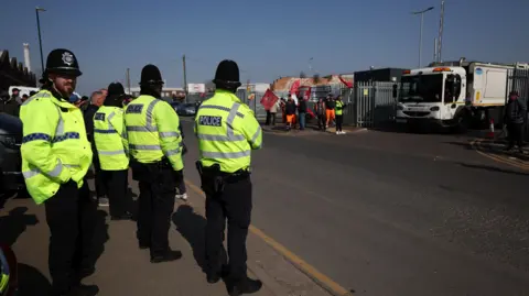 Police on a picket line at a Birmingham waste depot in April. They are wearing uniform and there is a bin lorry behind them as well as a couple of protestors. It is a bright and sunny day. 